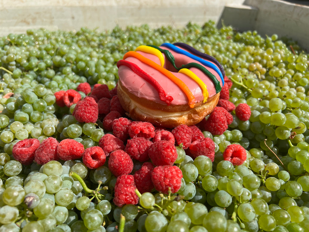 Colorful donut with pink icing and rainbow sprinkles surrounded by green grapes and red raspberries.